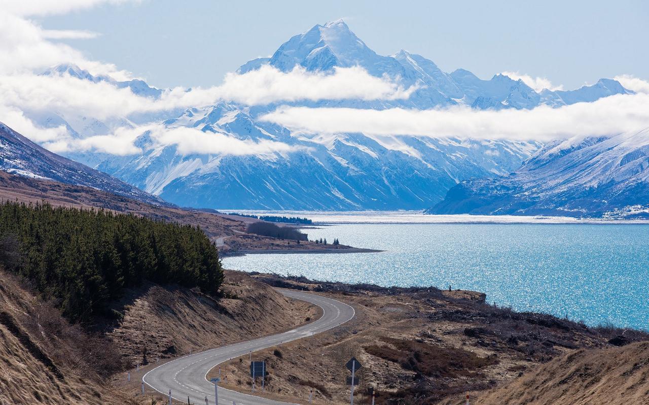 Winding road along Lake Pukaki with Mount Cook rising beyond the clouds