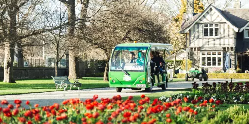 Electric shuttle passing colourful tulip beds and heritage buildings in Christchurch Botanic Gardens.