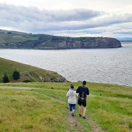 Two people walking down a grassy trail with coastal cliffs and Taylors Mistake in the distance