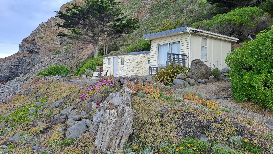 Whitewashed bach with gardens and ocean cliffs behind at Boulder Bay