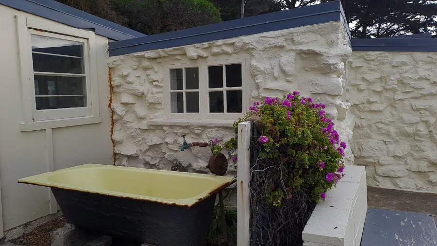 Old-fashioned outdoor bath and flower planter beside white stone bach at Boulder Bay