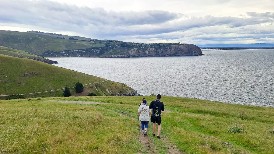 Two people walking down a grassy trail with coastal cliffs and Taylors Mistake in the distance