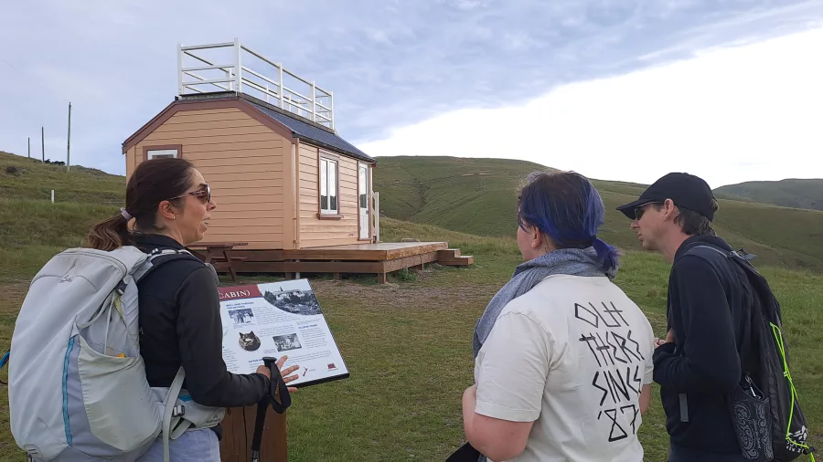 Crater Rim guide with backpack explaining Scott’s Cabin history to visitors near Godley Head