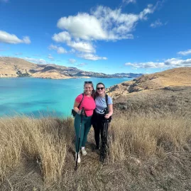 Two women with hiking poles smiling at a high vantage point above turquoise waters and rolling hills