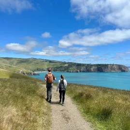 Two hikers walking along a grassy track with panoramic views toward Taylors Mistake and the Pacific Ocean