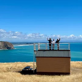 Three people waving from the rooftop deck of Scott’s Cabin above the Pacific