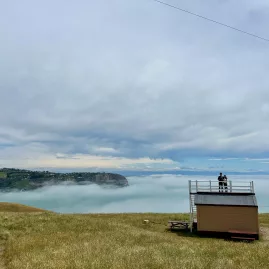 Person standing atop Scott’s Cabin roof above a cloud inversion at Godley Head