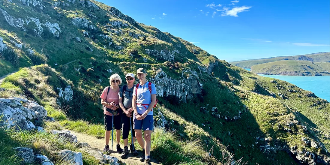 Three hikers standing on a narrow trail cut into the steep green cliffs above the sea on Banks Peninsula