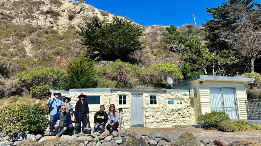 Group of hikers outside a traditional New Zealand bach with rocky cliffs and bright blue sky behind