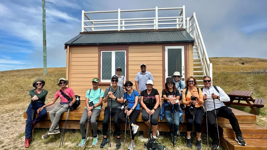 Group of hikers sitting in front of the Scott’s Cabin replica hut with walking poles and smiles