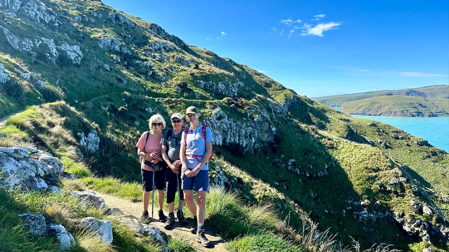 Three hikers standing on a narrow trail cut into the steep green cliffs above the sea on Banks Peninsula