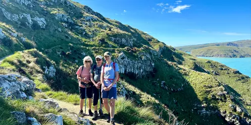 Three hikers standing on a narrow trail cut into the steep green cliffs above the sea on Banks Peninsula