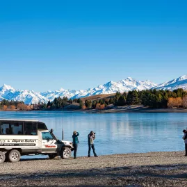 Alpine Safari vehicle parked by a lake with photographers capturing Aoraki/Mount Cook in the distance.
