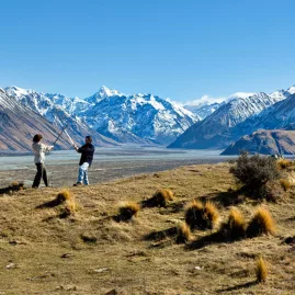 Two people pose with swords on a grassy ridgeline with snow-capped mountains behind.