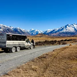 Tour vehicle parked on a gravel road with panoramic views of snow-covered peaks in the distance.