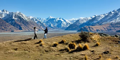 Two people pose with swords on a grassy ridgeline with snow-capped mountains behind.