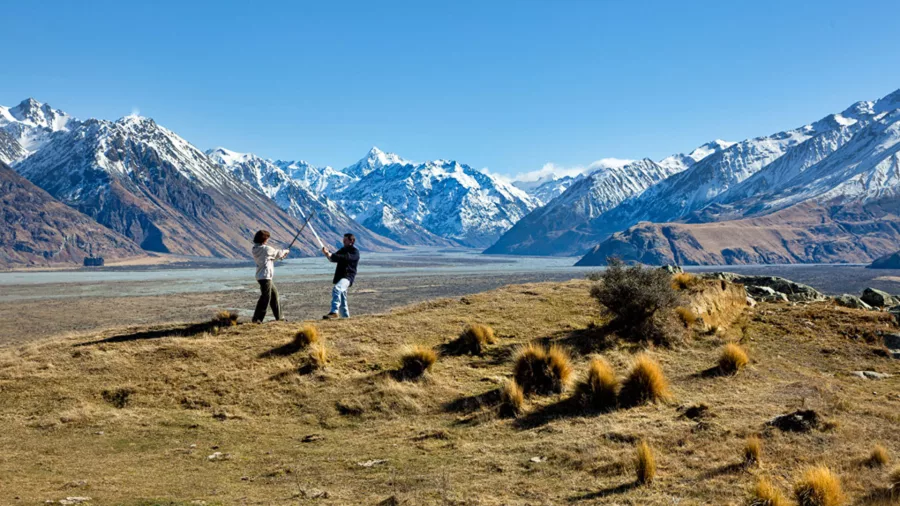 Two people pose with swords on a grassy ridgeline with snow-capped mountains behind.