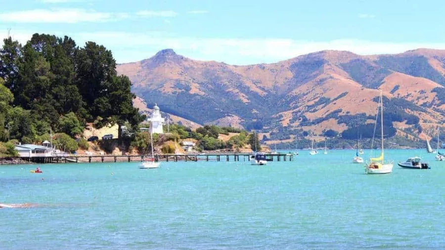 Boats anchored near Akaroa Lighthouse with pine trees and mountains in the background