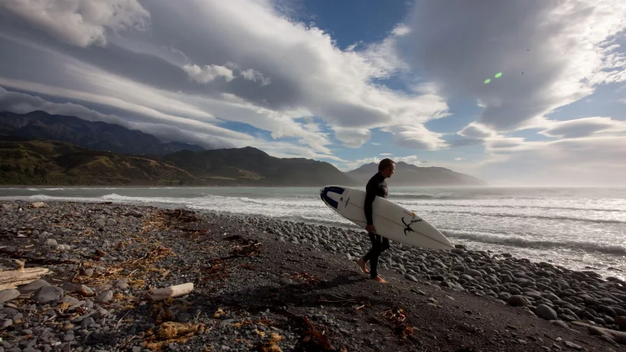 Surfer walking along Kaikōura’s rugged beach at sunset