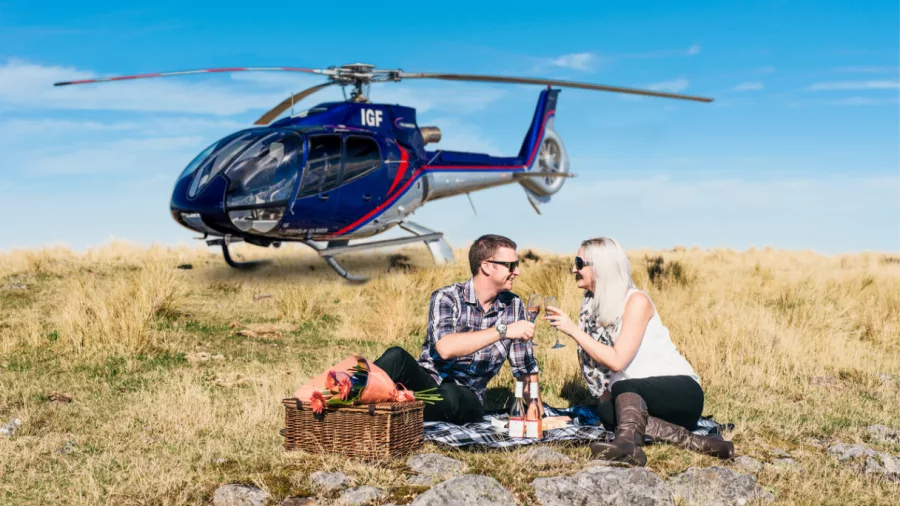 Couple enjoying picnic together with helicopter landed behind in a grassy alpine setting