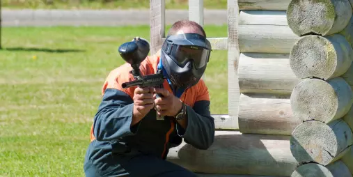Paintball player in orange and black gear aiming behind a wooden wall at Hanmer Springs