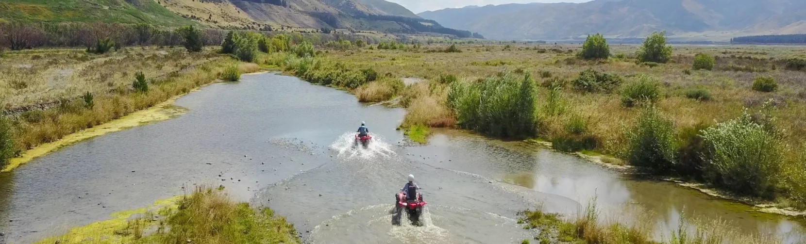 Two quad bikes splashing through a wetland trail in Hanmer Springs with dramatic mountains in the background