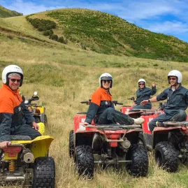Smiling group in orange vests posing on quad bikes on a grassy hillside in Hanmer Springs