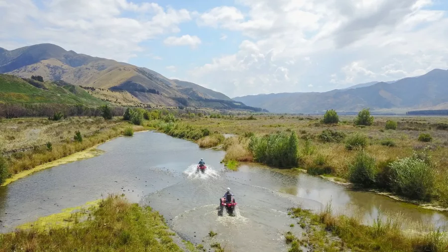 Two quad bikes splashing through a wetland trail in Hanmer Springs with dramatic mountains in the background