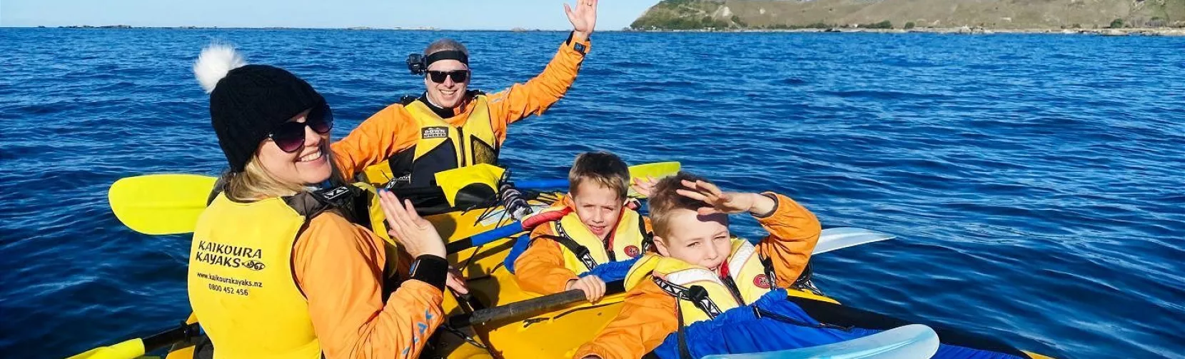Mum and kids waving from yellow kayaks on calm blue water during a Kaikōura tour