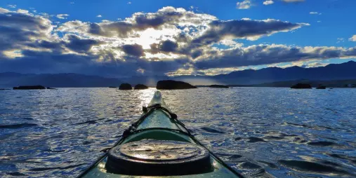 Sea kayak gliding across calm waters in Kaikōura at sunset, with dramatic clouds and distant mountains on the horizon.