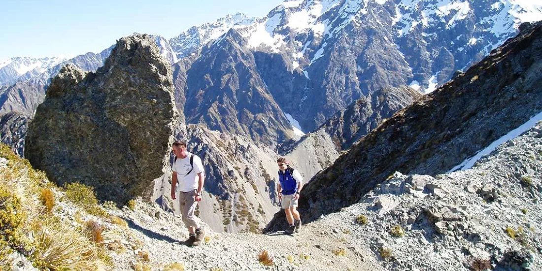 Two hikers making their way up the rocky trail on Mount Sebastopol with snow-covered peaks in the background.