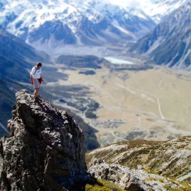 Hiker standing on a rocky outcrop on Mount Sebastopol overlooking the Aoraki/Mount Cook village and valley.