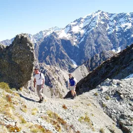 Two hikers making their way up the rocky trail on Mount Sebastopol with snow-covered peaks in the background.