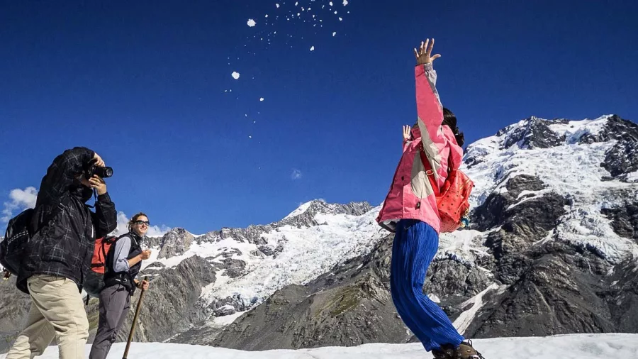 Person tossing snow or ice into the air while others look on, with Tasman Glacier mountains in the background.