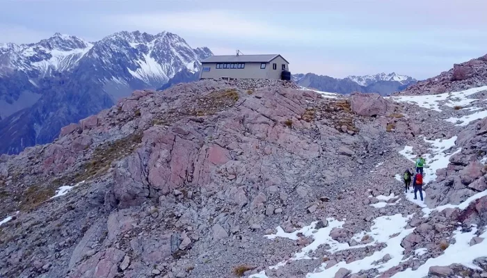 Hikers nearing Caroline Hut on a rocky trail surrounded by snow-dusted peaks.