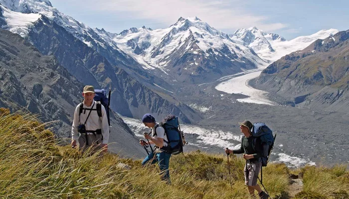 Three hikers climb a grassy ridge with views over the Tasman Glacier in Aoraki Mt Cook National Park.