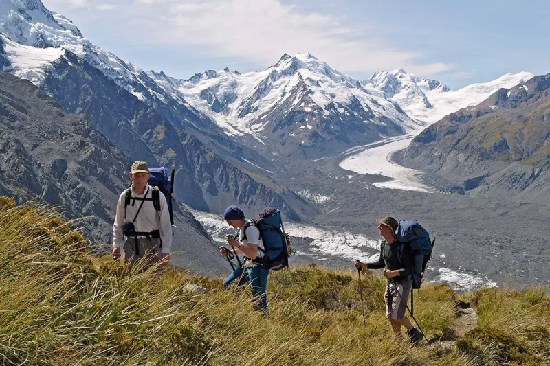 Three hikers climb a grassy ridge with views over the Tasman Glacier in Aoraki Mt Cook National Park.