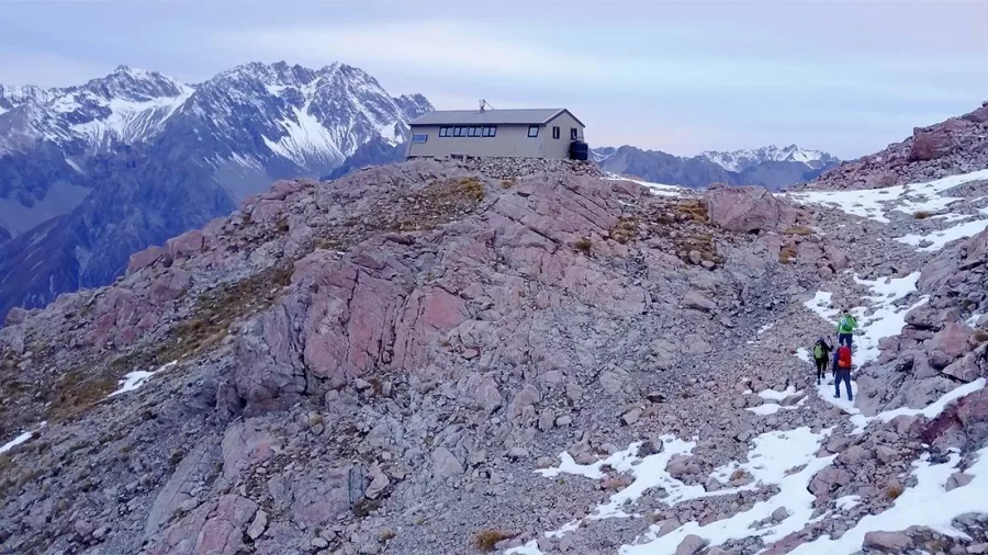 Hikers nearing Caroline Hut on a rocky trail surrounded by snow-dusted peaks.