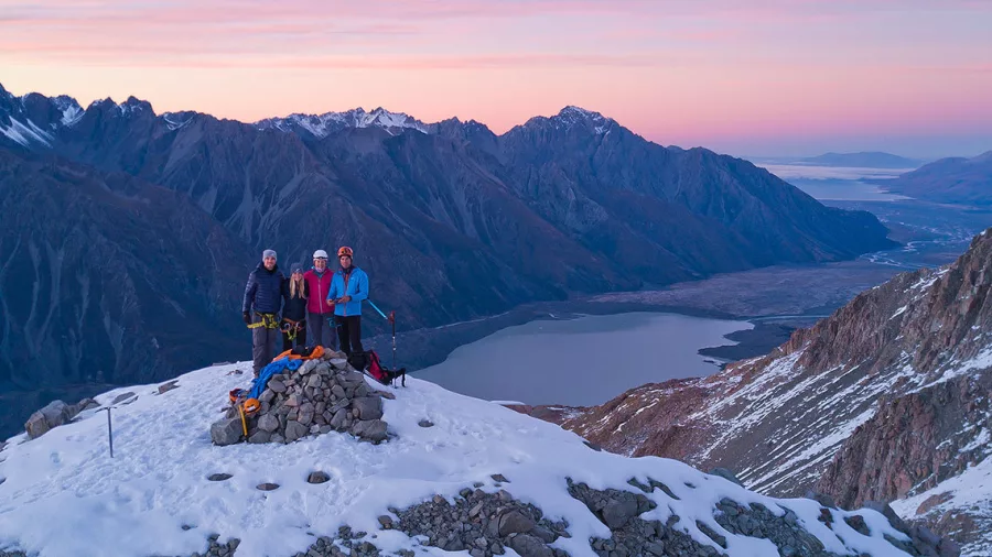 Hikers stand beside a summit cairn with Lake Pukaki visible in the distance during sunrise.