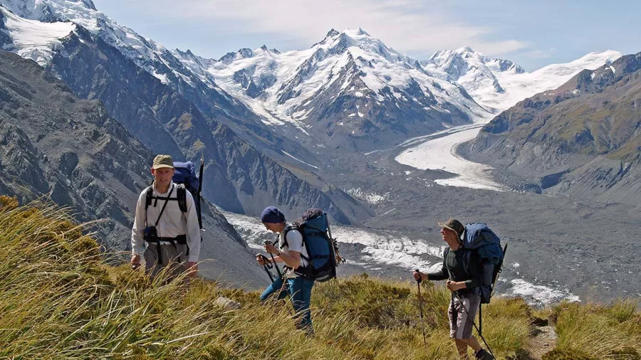 Three hikers climb a grassy ridge with views over the Tasman Glacier in Aoraki Mt Cook National Park.