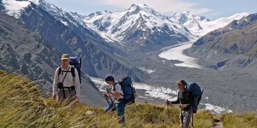 Three hikers climb a grassy ridge with views over the Tasman Glacier in Aoraki Mt Cook National Park.