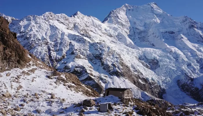 Snow-covered Caroline Hut with towering Aoraki Mount Cook behind.
