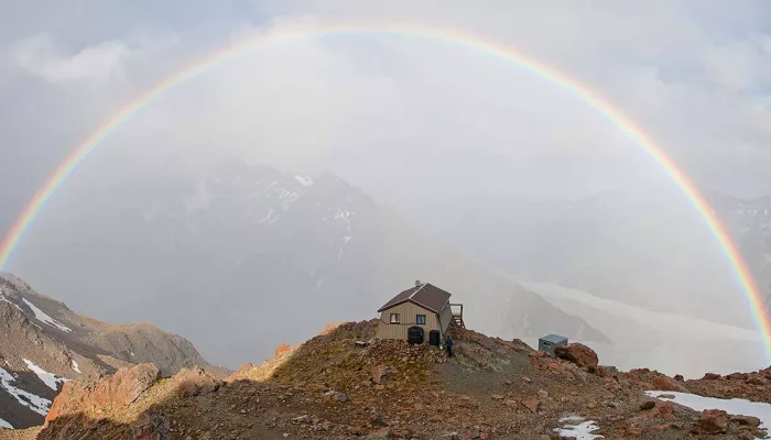A full rainbow arcs over Caroline Hut on a dramatic weather day in Mount Cook National Park.
