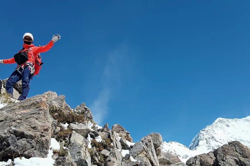 Climber waving from a rocky outcrop with Aoraki Mount Cook in the distance on Ball Ridge.
