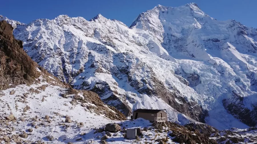 Snow-covered Caroline Hut with towering Aoraki Mount Cook behind.