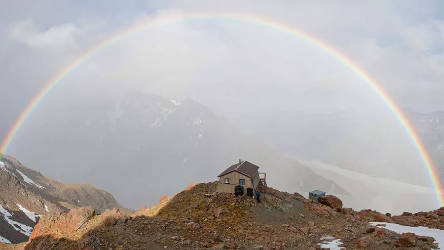 A full rainbow arcs over Caroline Hut on a dramatic weather day in Mount Cook National Park.