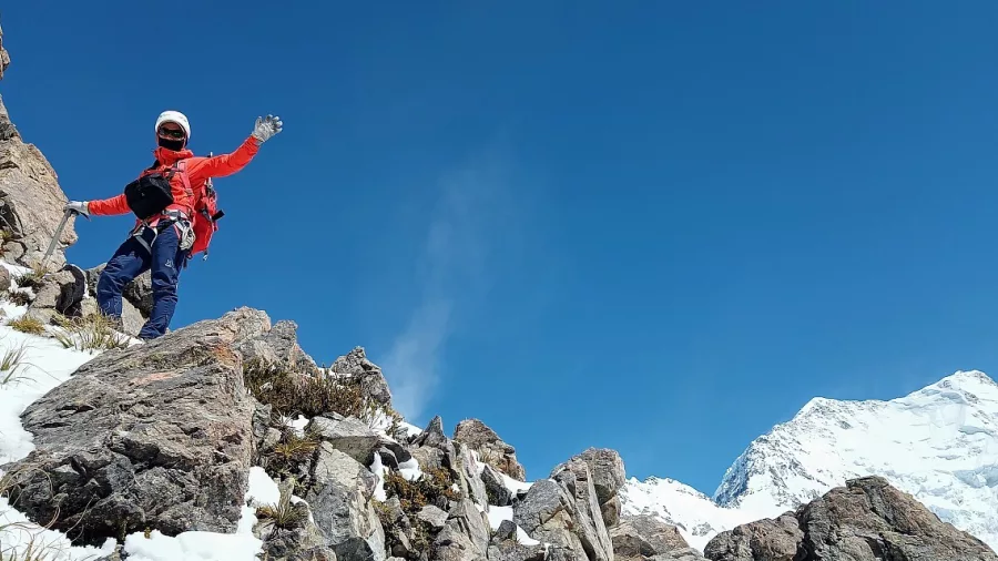 Climber waving from a rocky outcrop with Aoraki Mount Cook in the distance on Ball Ridge.
