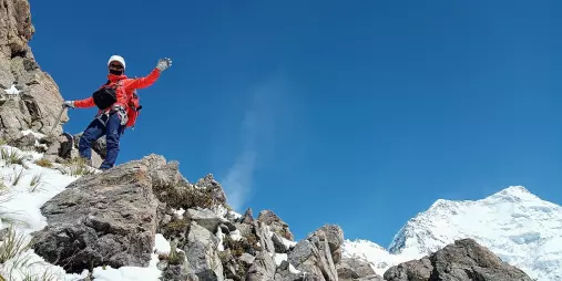 Climber waving from a rocky outcrop with Aoraki Mount Cook in the distance on Ball Ridge.