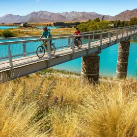 Cyclists riding across a bridge at Lake Tekapo with the Church of the Good Shepherd in the background