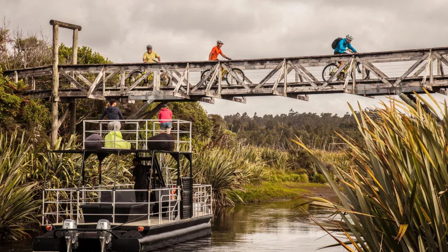 Cyclists riding over a wooden bridge above a boat on Lake Mahinapua near Hokitika